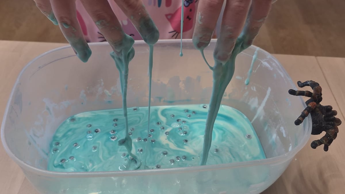 Hands lifting long drips of blue oobleck from a tub dotted with googly eyes, with a plastic tarantula on the rim