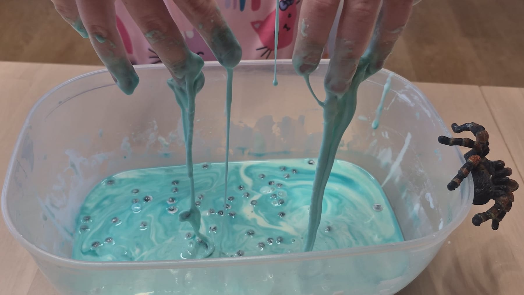 Hands lifting long drips of blue oobleck from a tub dotted with googly eyes, with a plastic tarantula on the rim