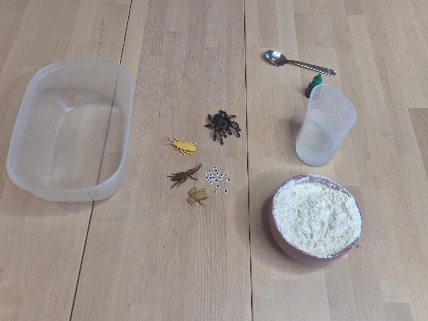 Oobleck materials on a wooden table: an empty plastic tub, a bowl of cornstarch, a jug of water, green food colouring, a spoon, googly eyes and plastic bugs