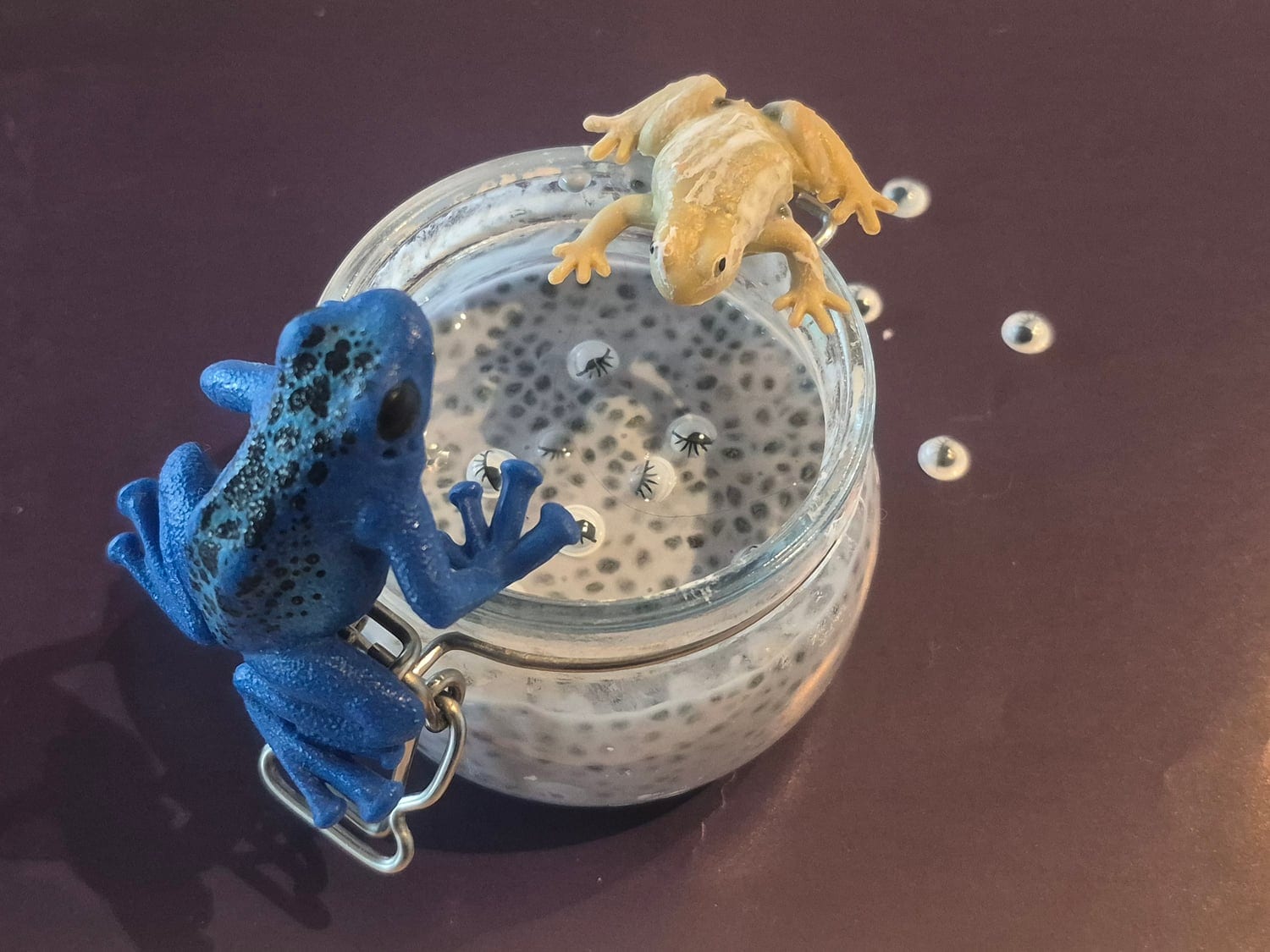 A glass jar of chia seed slime that looks like frog spawn, with a blue and a yellow toy frog climbing over it and googly eyes scattered around