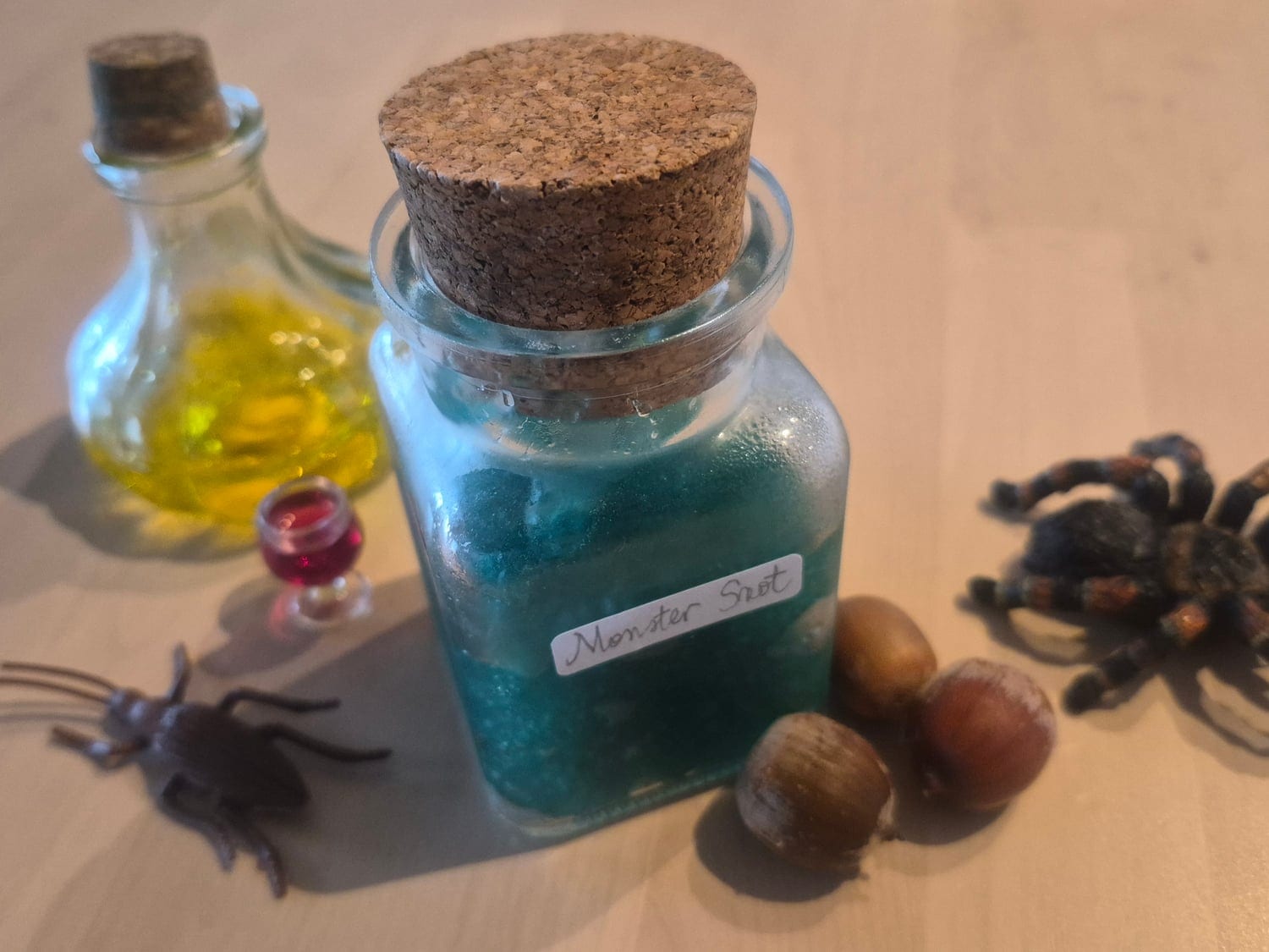 Green monster snot in a corked glass jar with a handwritten label, surrounded by plastic spiders, a tarantula, chestnuts and a little potion bottle