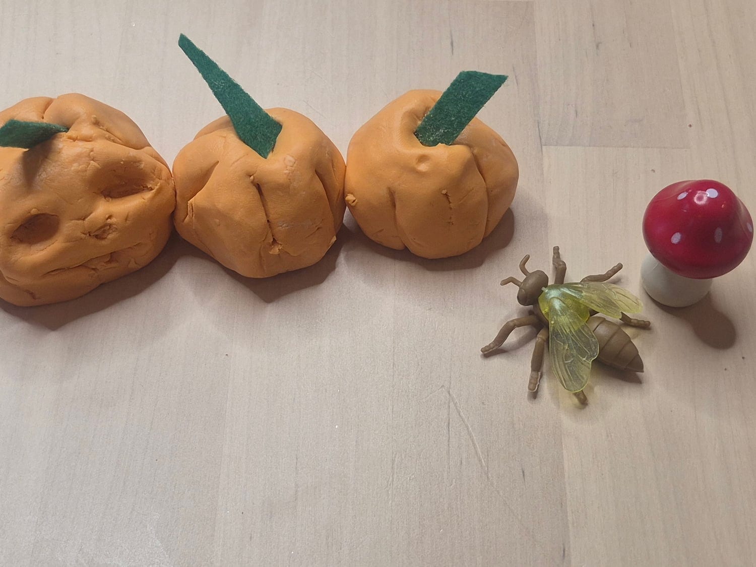 Three orange pumpkins shaped from cornstarch slime with green paper stems, next to a toy fly and a toy mushroom